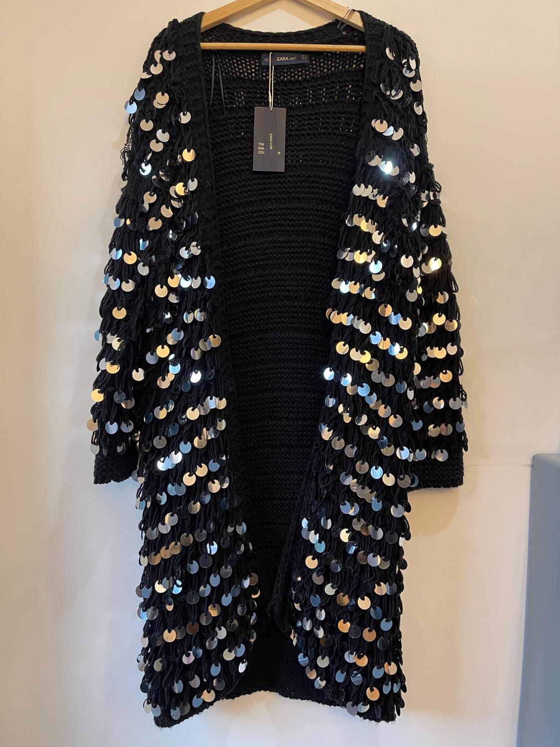 Black knitted jacket with sparkly sequins, hanging on a wooden hanger. Visible "ZARA" label.