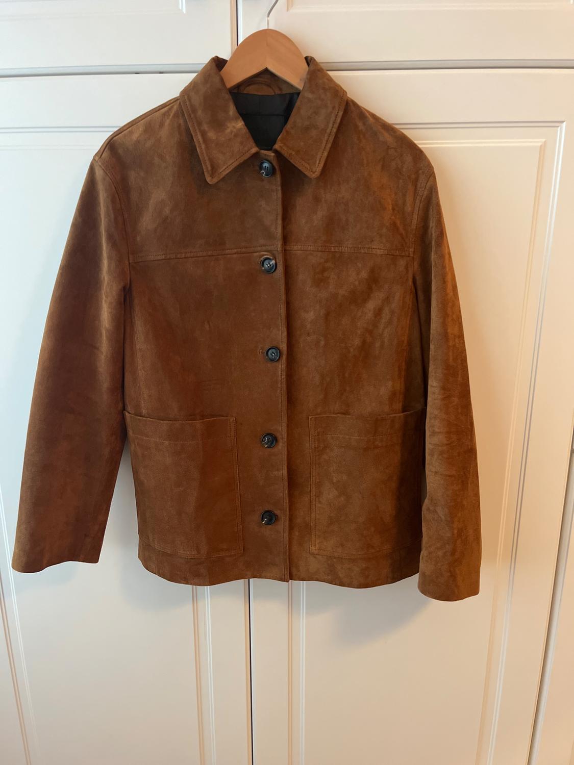 Brown suede jacket hanging on a wooden hanger, with dark buttons and front pockets, against a background of white doors.