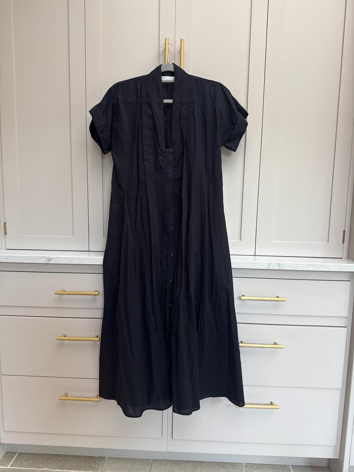 Short-sleeved black dress with front buttons, hanging on a clothes hanger in front of a light-coloured chest of drawers with golden handles.