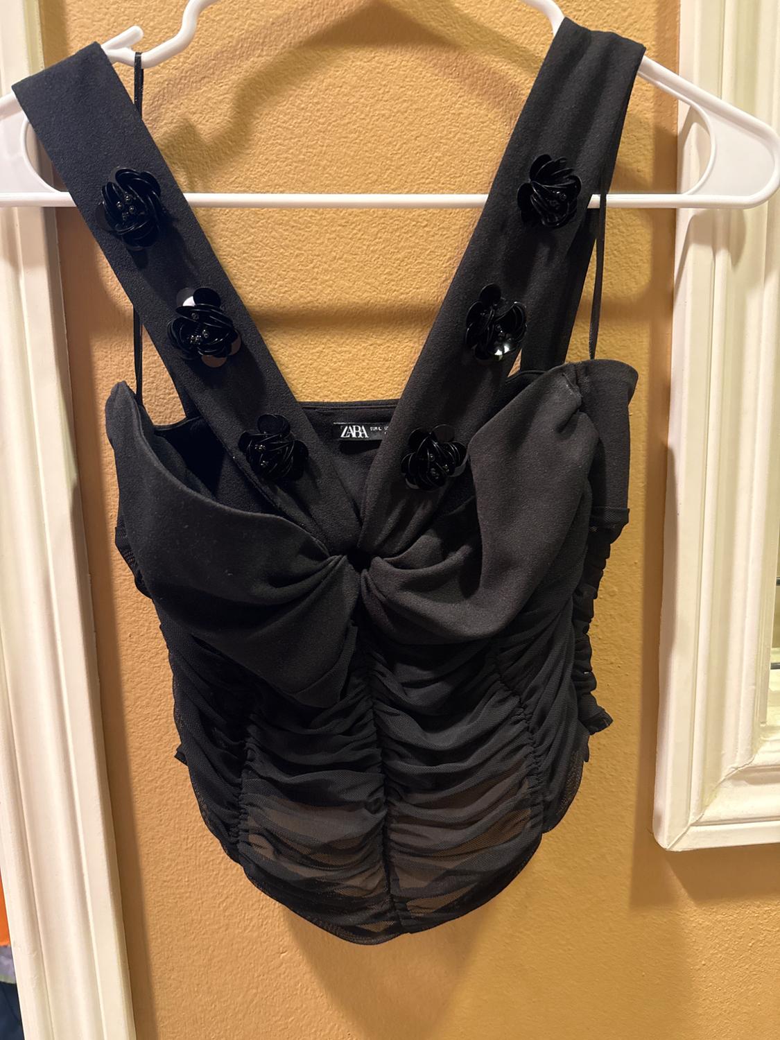 Black top with wide straps decorated with black flowers, bow detail in the front and ruched design, hanging on a white hanger.