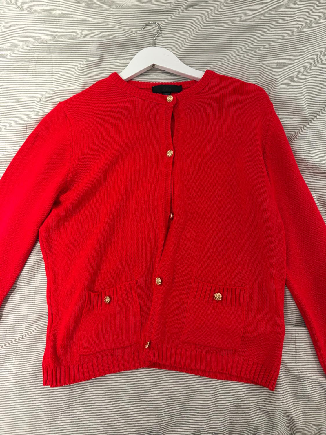 Red cardigan with gold buttons and two front pockets, hanging on a white hanger over grey and white striped fabric.