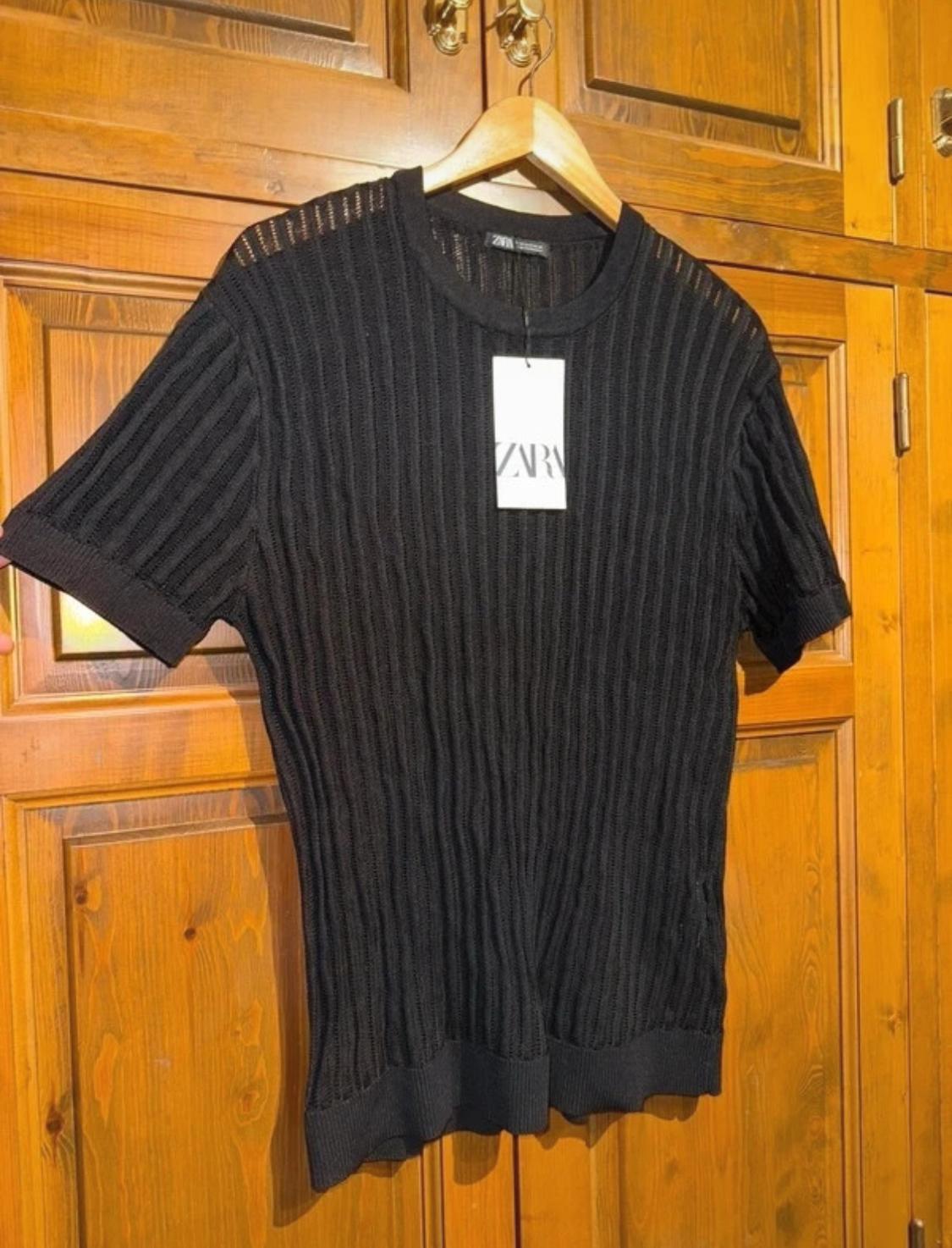 Black knitted T-shirt with ribbed texture, hanging on a wooden hanger in front of a brown wardrobe. Visible label: ZARA.
