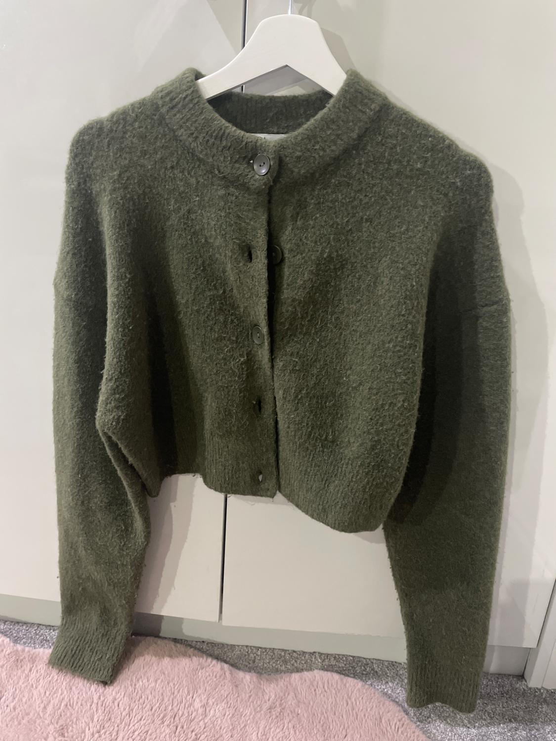 Olive green wool cardigan with buttons, hanging on a white hanger against a light background, on a pink rug.