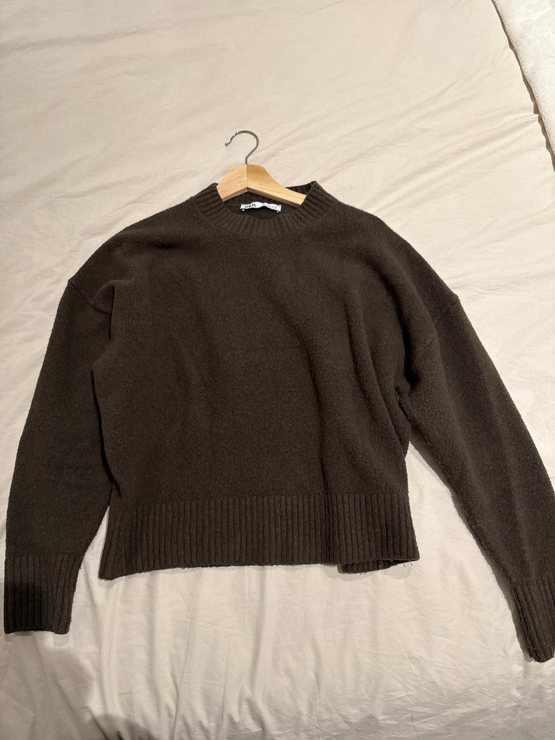Dark brown chunky knit crew-neck jumper, placed on a beige bed. It's hanging on a wooden hanger.