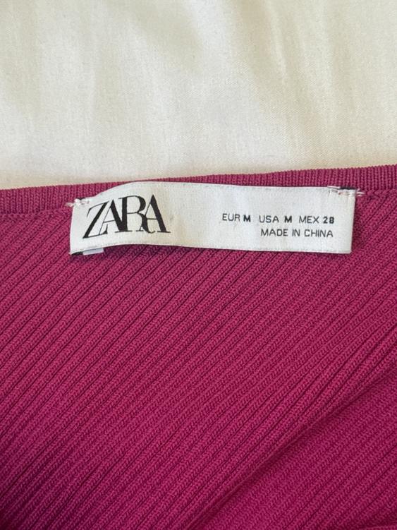 ZARA clothing tag on pink fabric. Text: "EUR M USA M MEX 28 MADE IN CHINA". White background.