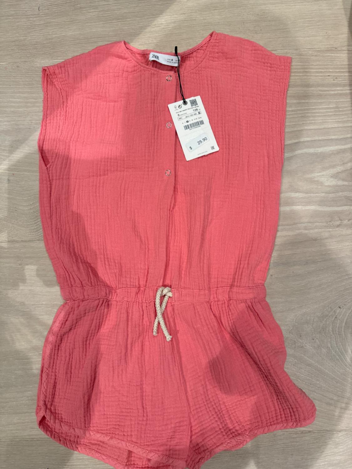 Pink sleeveless jumpsuit with crinkled texture, drawstring waist, and a $29.90 price tag on a light wooden background.