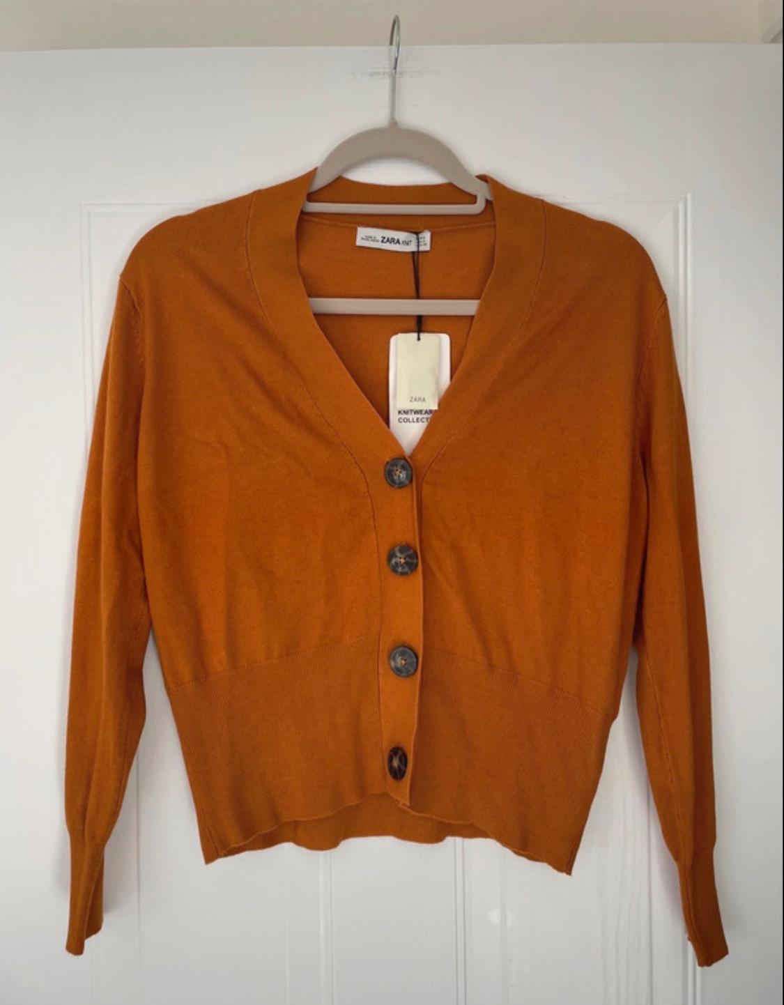 Orange V-neck button-up cardigan hanging on a white door. Text: ZARA KNIT, ZARA KNITWEAR COLLECT.