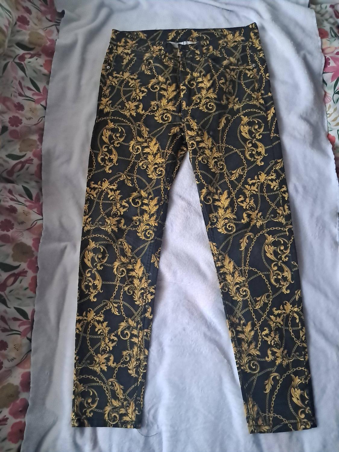 Black trousers with a gold baroque print of chains and leaves, laid out on a light-coloured fabric with a floral background.