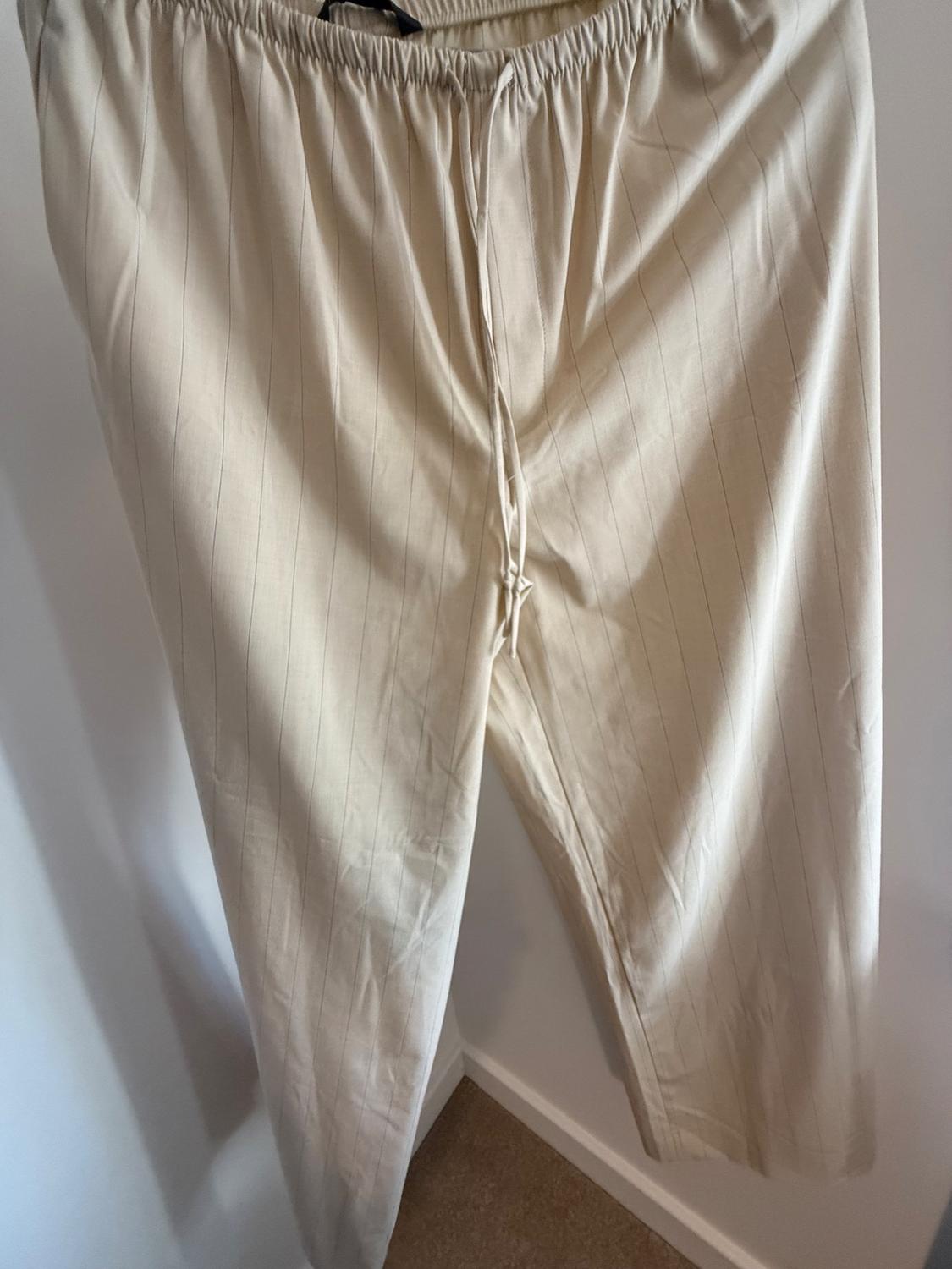 Beige fine-striped trousers with an elasticated waistband and drawstring, hanging against a white wall in a relaxed setting.