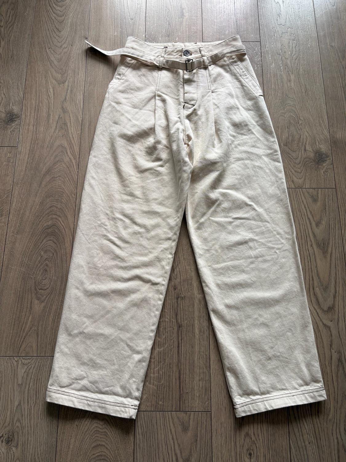 Beige wide-leg trousers with pleats and a matching belt, laid out on a wooden floor. Relaxed, casual-style composition.