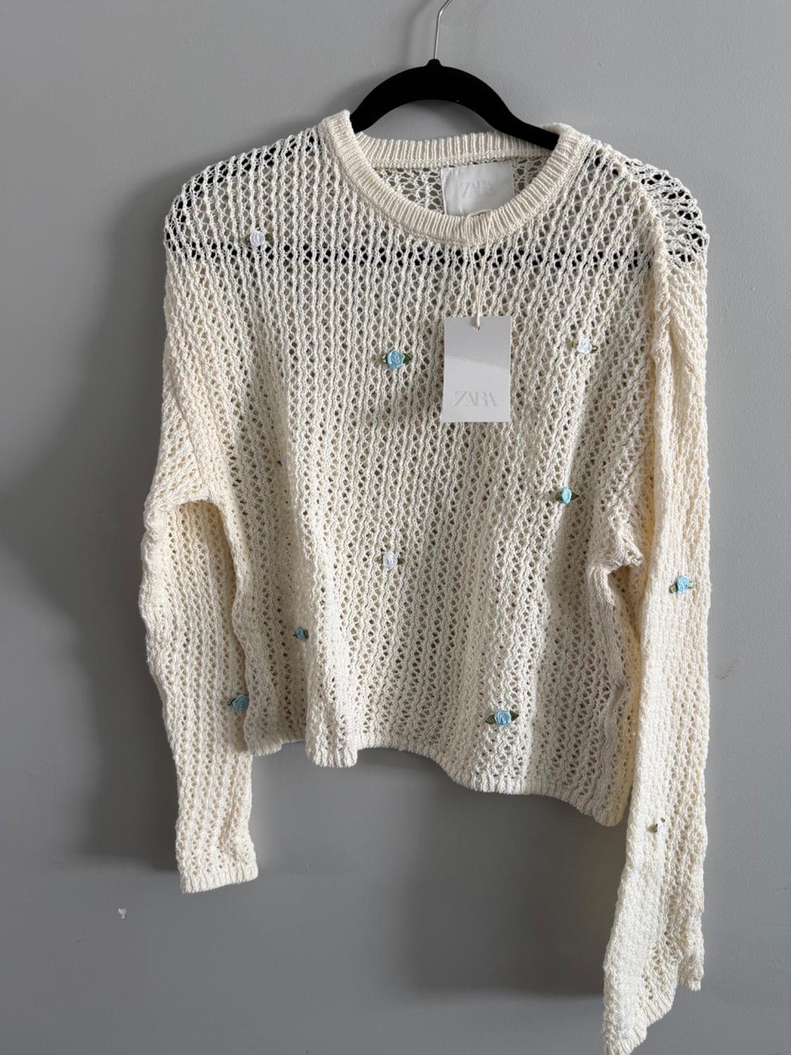White open-knit sweater with blue and white floral details, hanging on a black hanger. Visible tag with text "ZARA".