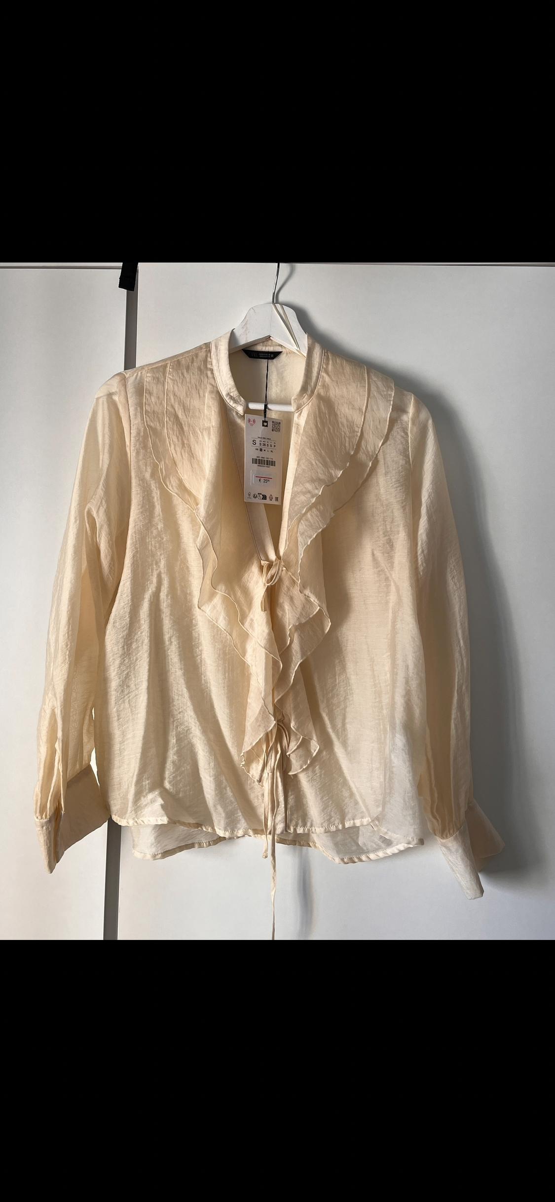 Beige frilled blouse hanging on a white hanger. Visible label with price details and size S. Black and white background.