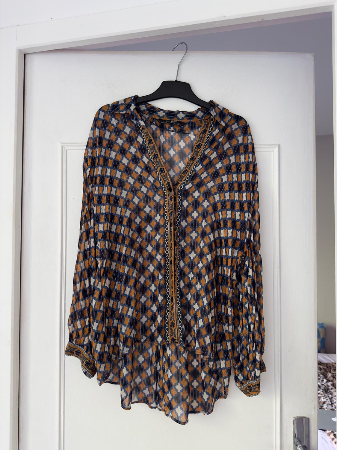 A chiffon blouse with a geometric print in shades of brown, blue and white, hanging on a hanger over a white door.