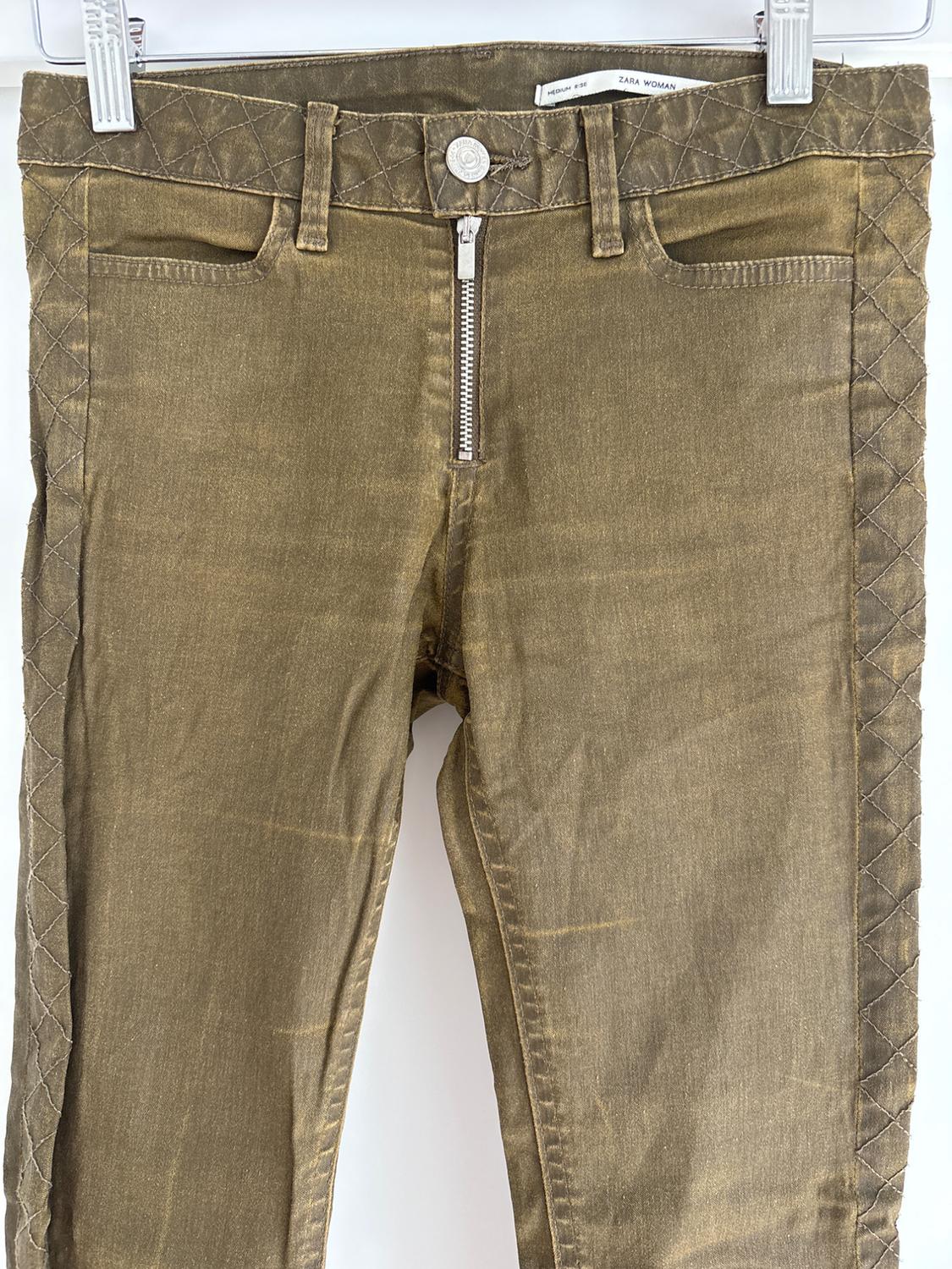 Brown denim trousers with a front zip and diamond-stitched detailing on the sides. "ZARA WOMAN" label visible at the waist.