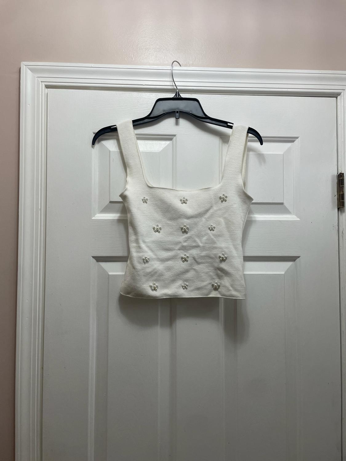White knit top with a square neckline and flower-shaped pearl details, hanging on a hanger on a white door.