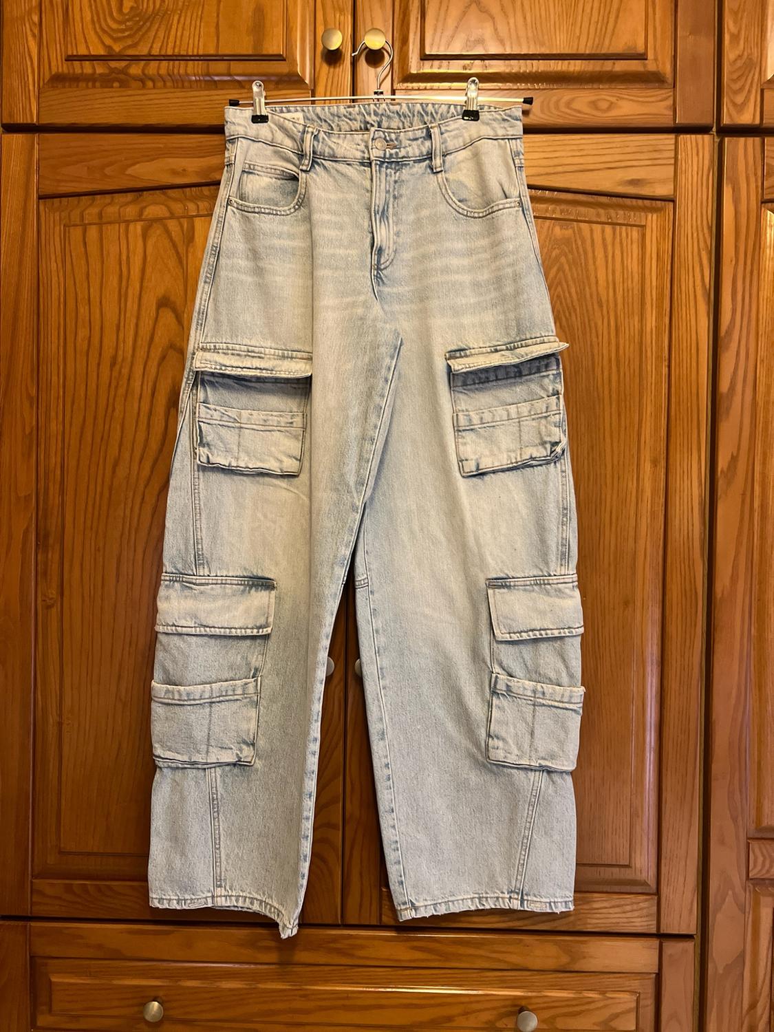 Light blue cargo jeans with multiple pockets, hanging on a hanger in front of a wooden wardrobe.