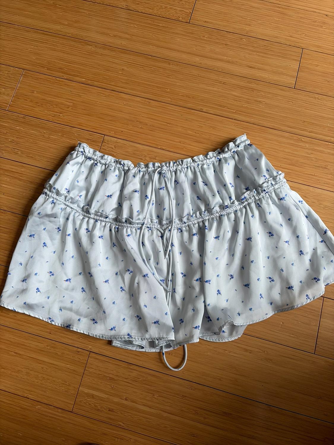 Light blue short skirt with a blue floral print, elastic waist, drawstring, and ruffles, on a wooden floor.