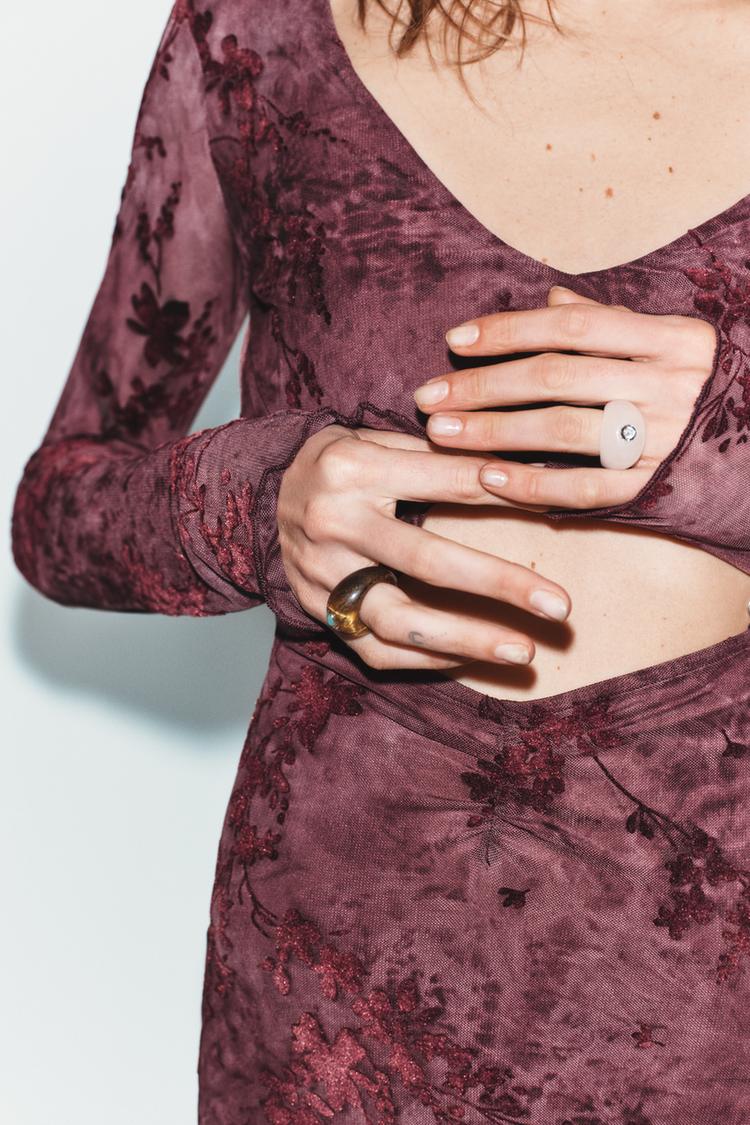 Detailní záběr rukou se dvěma prsteny, jedním tmavým a druhým světlým s leskem, na vínových šatech s květinovým potiskem.