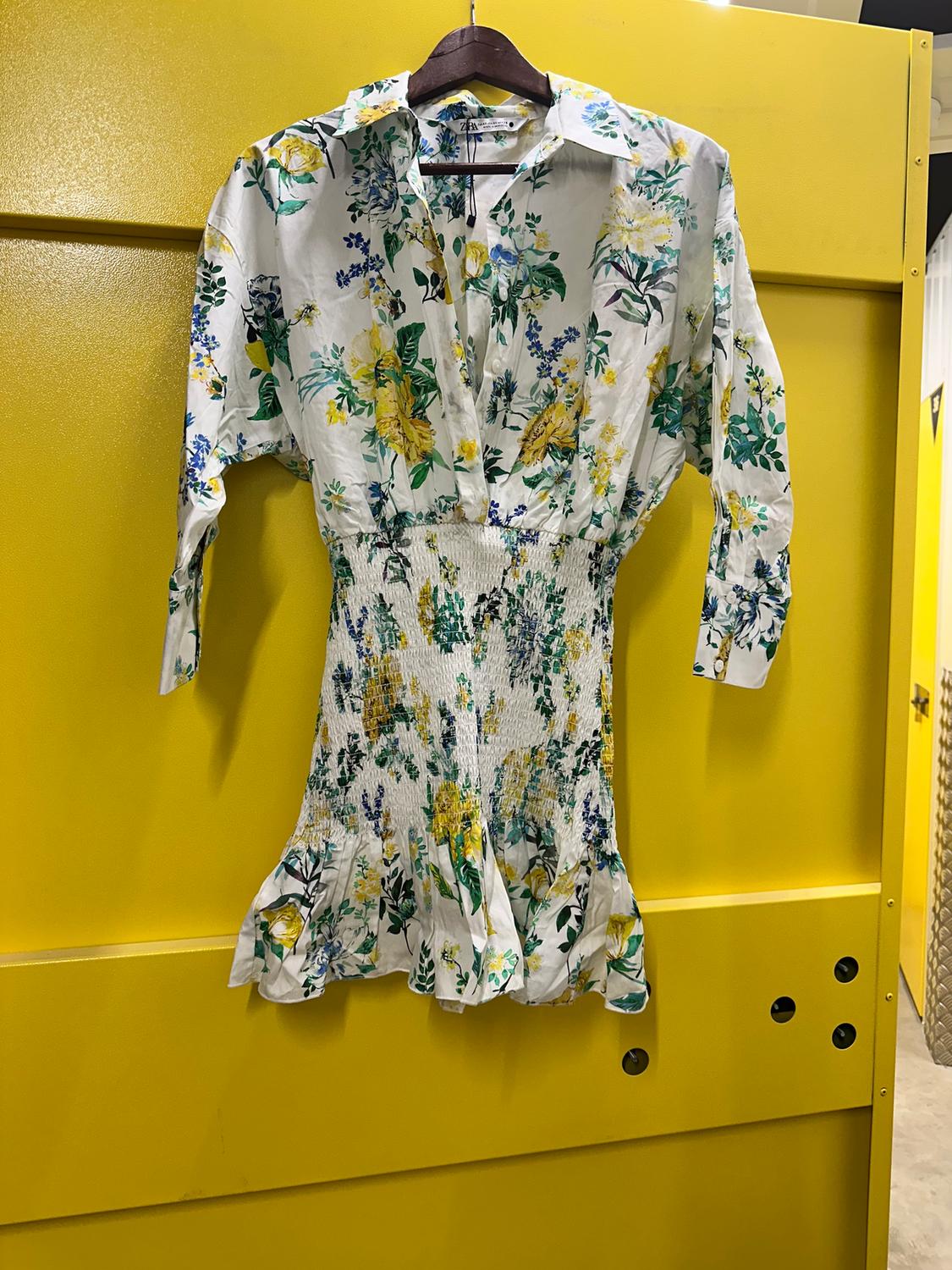 Floral dress with a white background and yellow and blue floral print, hanging on a hanger against a bright yellow wall.