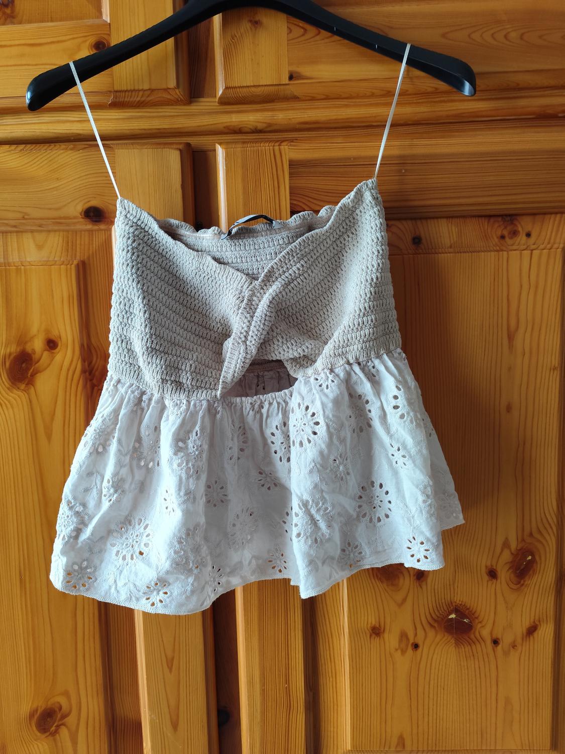 Black hanger vest top, with a beige knit upper and a white lace lower with floral embroidery, wooden background.