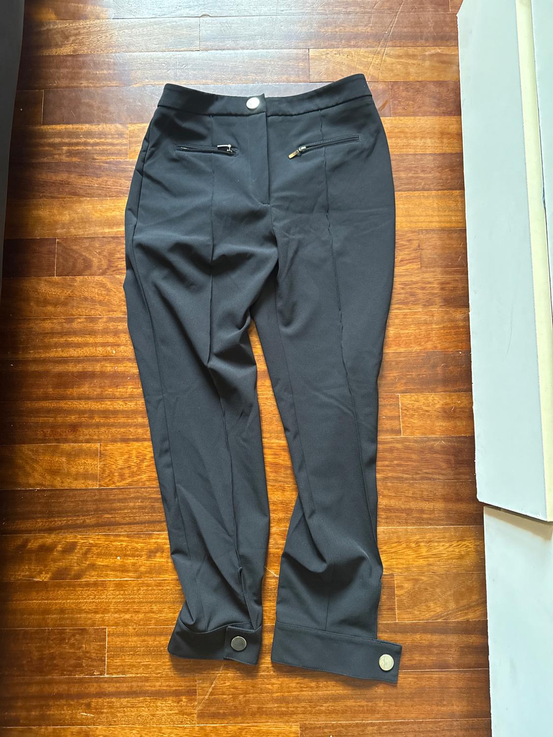 Black trousers with zip pockets and ankle buttons, on a brown wooden floor.