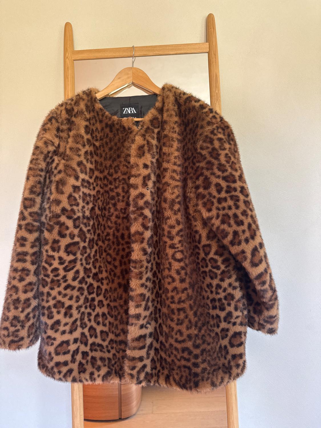 Faux fur leopard print coat hanging on a wooden hanger in front of a mirror. Visible tag: ZARA.