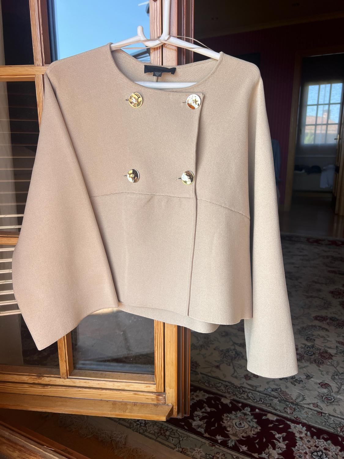 Beige wide-sleeve jacket with gold buttons hanging on a wooden window. Against the background, a room.