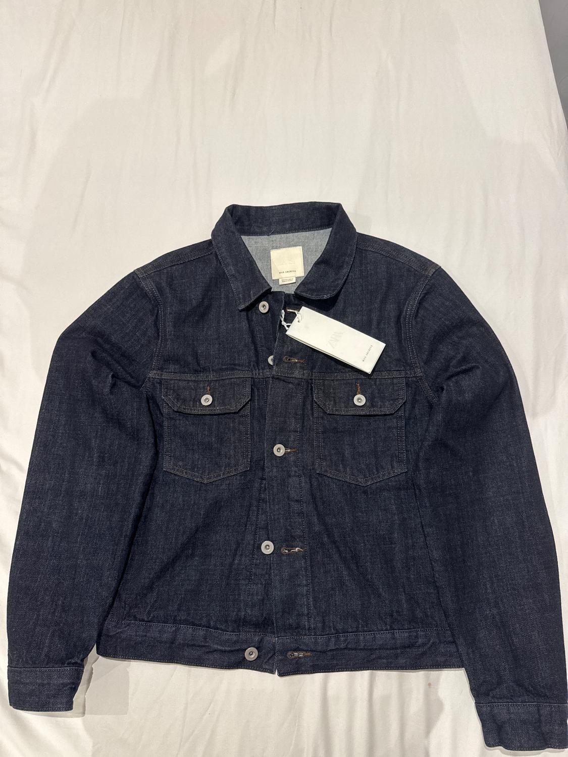 Dark blue denim jacket with front buttons and two chest pockets, on a white surface. Visible tag.