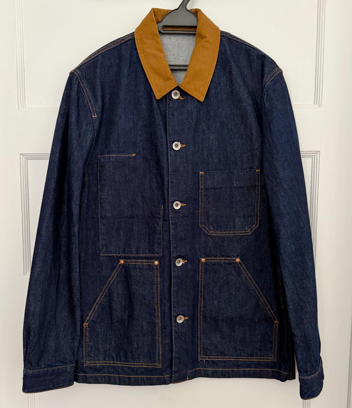 Dark blue denim jacket with a brown collar and patch pockets, hanging on a black hanger over a white door.
