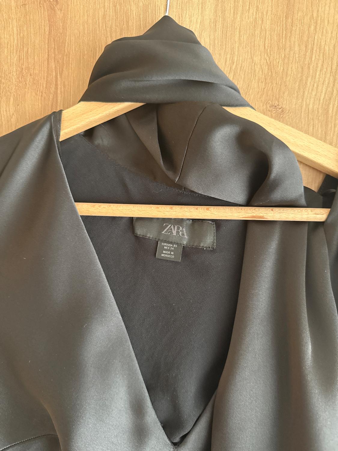 Black satin shirt hanging on a wooden hanger, with a visible label that reads "ZARA" and "MADE IN MOROCCO". Light wooden background.