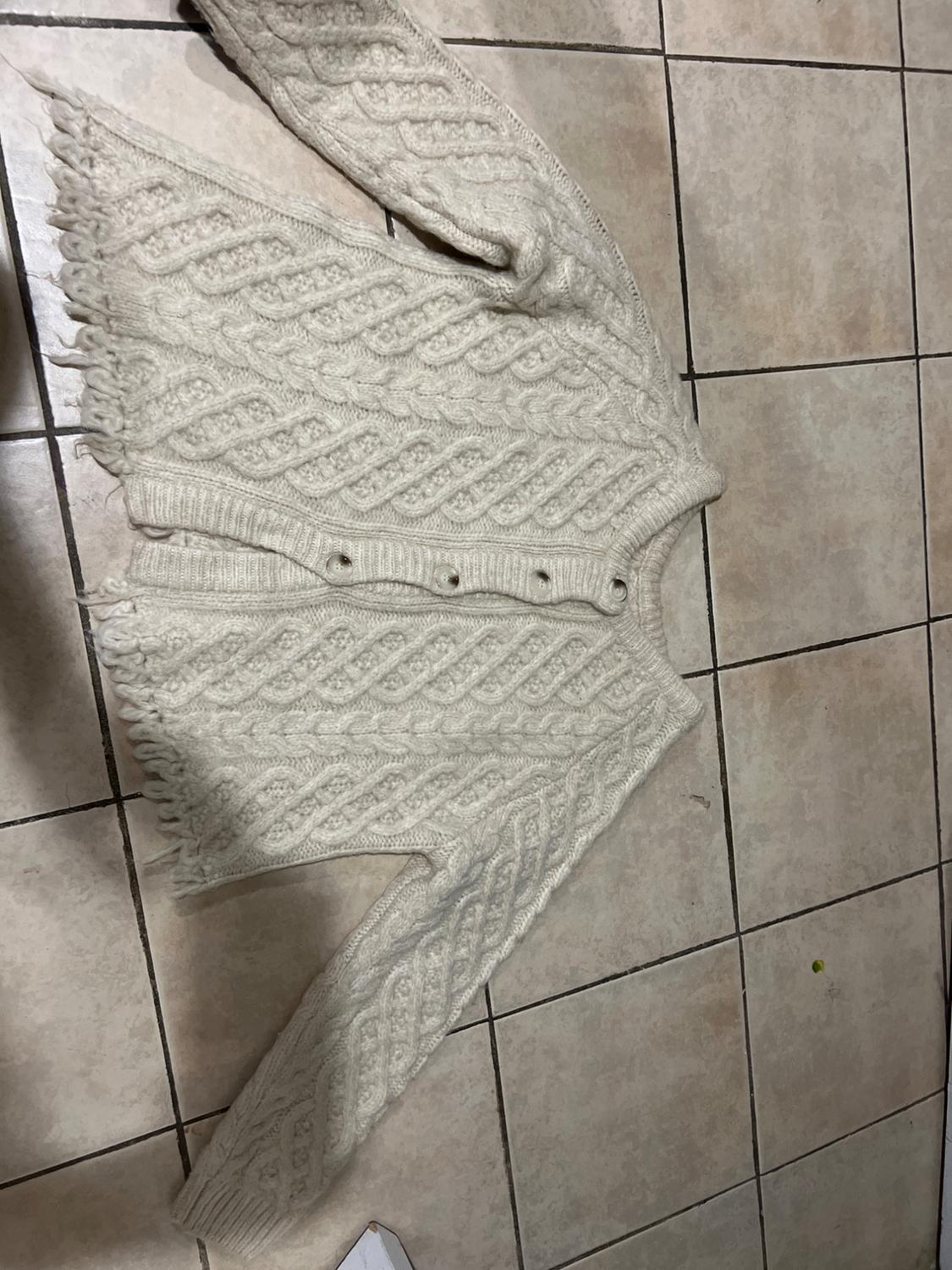 Cream knitted jumper with cable knit design and fringing on the edges, placed on a beige tiled floor.