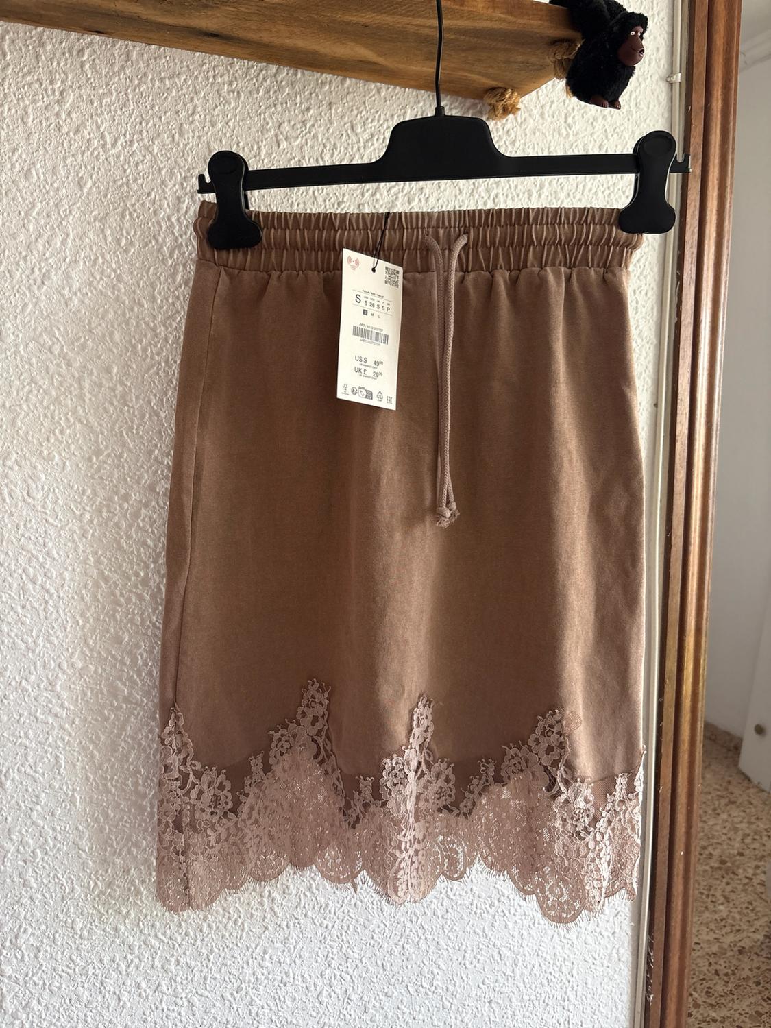Brown skirt with elasticated waist, drawstring and floral lace hem. Hanging from a black hanger with a tag in front of a textured white wall.