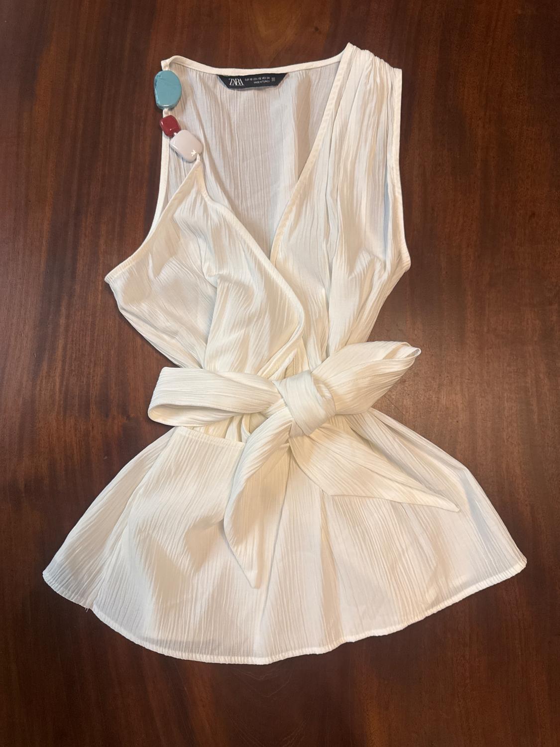 White sleeveless blouse with a pleated texture and a knotted belt, on a wooden background. Coloured bead detail on the strap.