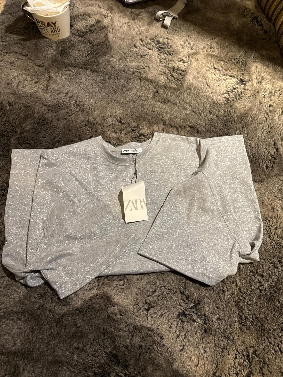 Shiny gray T-shirt folded on a shag rug with a ZARA tag. Mug in the background with the text PRAY HOPE AND don't worry.