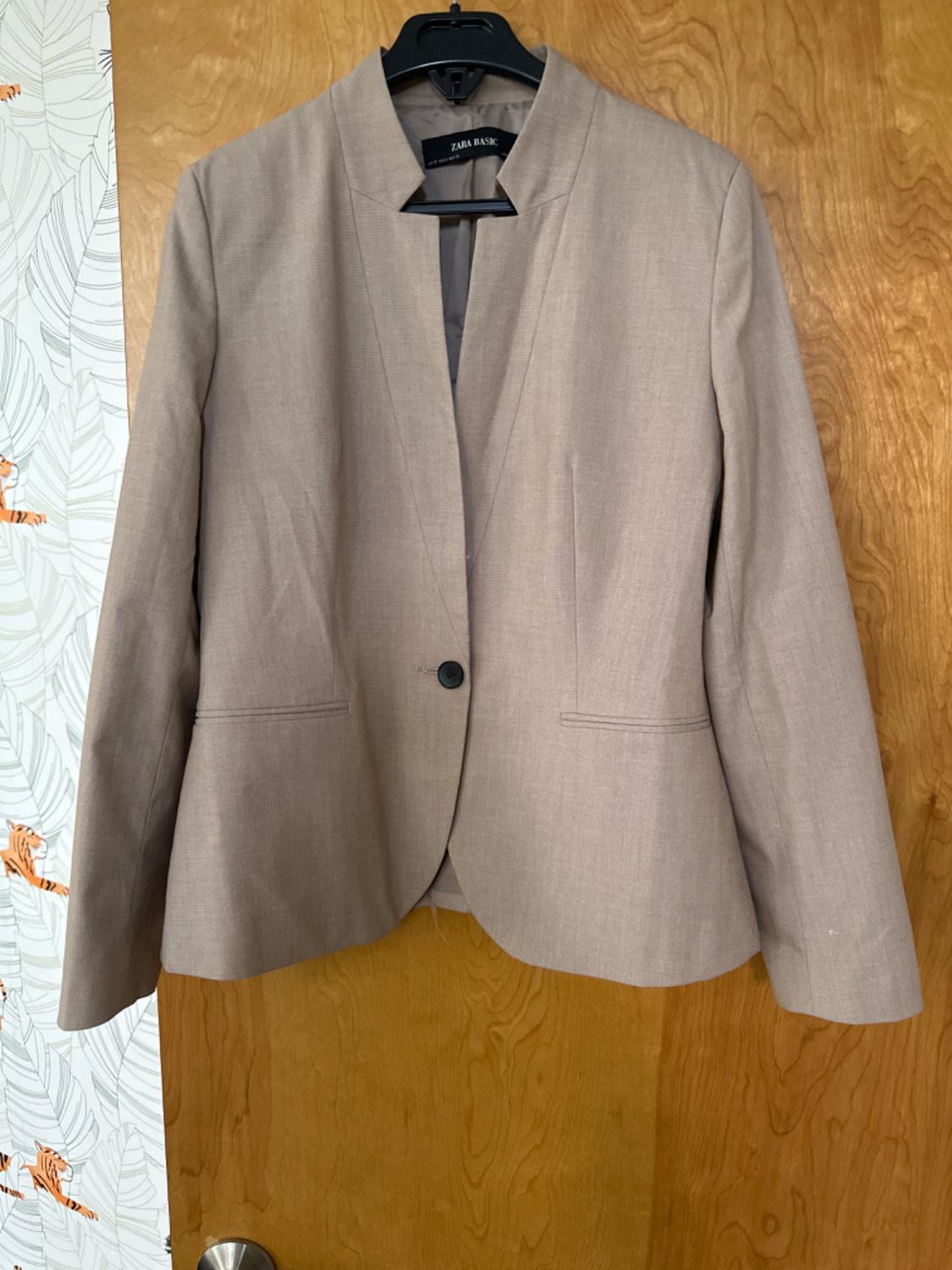 Beige Zara Basic jacket with an open collar and a single central button, hanging on a coat hanger in front of a wooden door. Background with a leaf and tiger design.