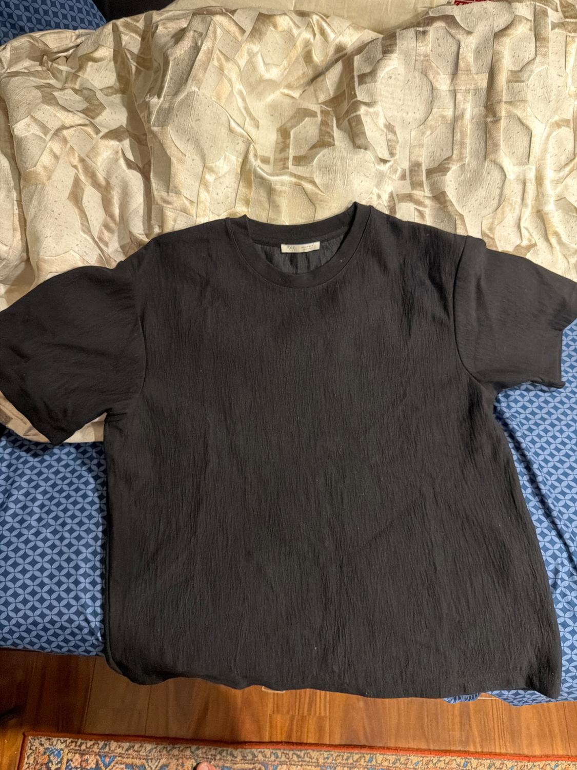 Black short-sleeved t-shirt in a textured fabric, laid out on a bed with blue and beige patterned sheets.