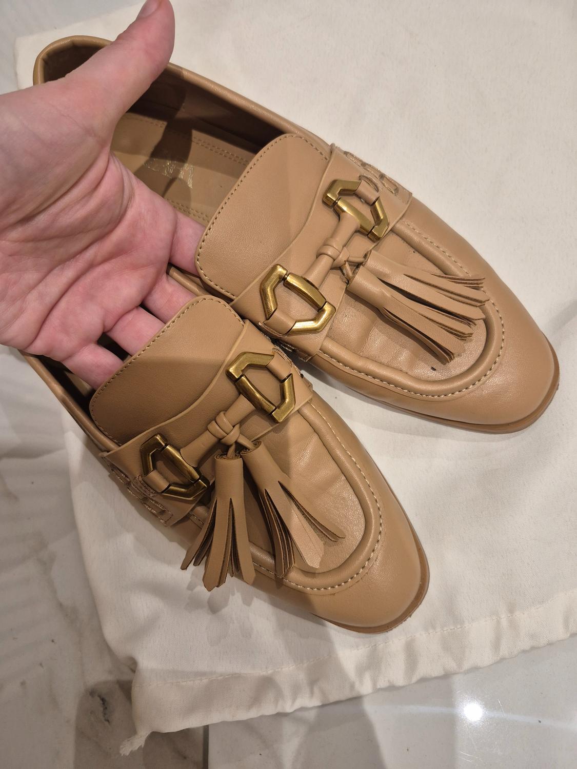 A pair of beige loafers with tassels and gold buckles, held by a hand over a white fabric.