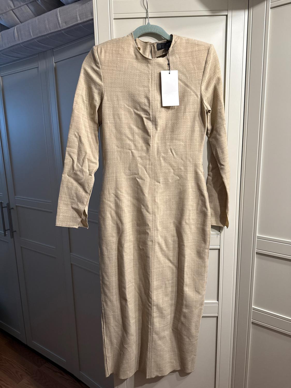 Long-sleeved beige dress hanging on a blue hanger, with a white tag, in front of a white wardrobe.