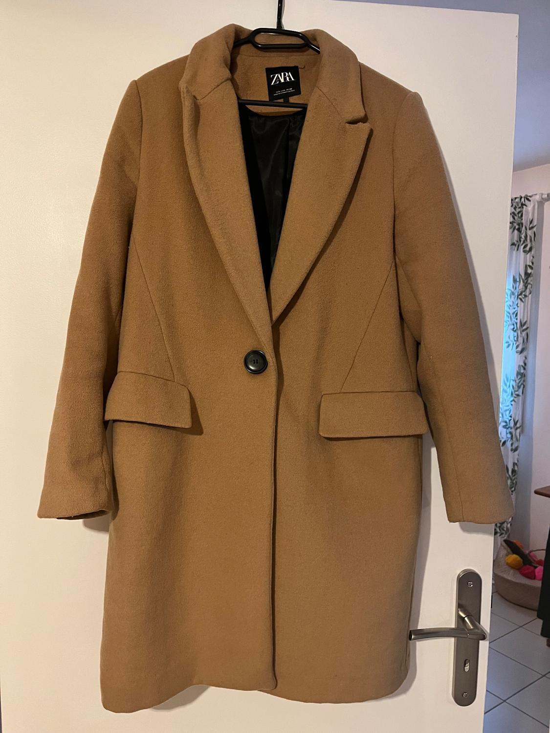 Camel coat hanging on a white door, with a lapel and a black button. Visible label with the text "ZARA". Interior background of a room.