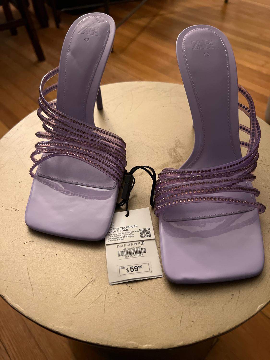 Lilac high-heeled sandals with decorative straps and a $59.90 price tag on a light-coloured round wooden table.
