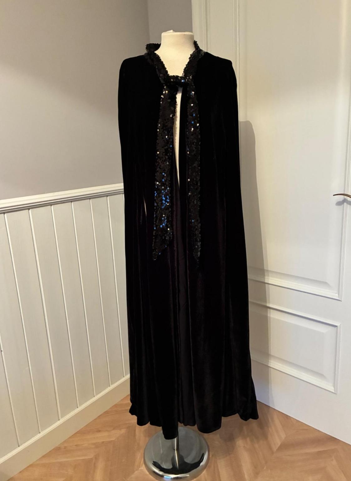 Long black velvet cloak with sequinned straps at the neck, displayed on a mannequin in a room with a wooden floor.