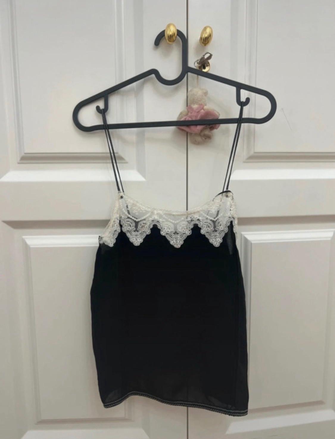 Black camisole top with spaghetti straps and white lace trim at the neckline, hanging on a hanger over a white door with a teddy bear.