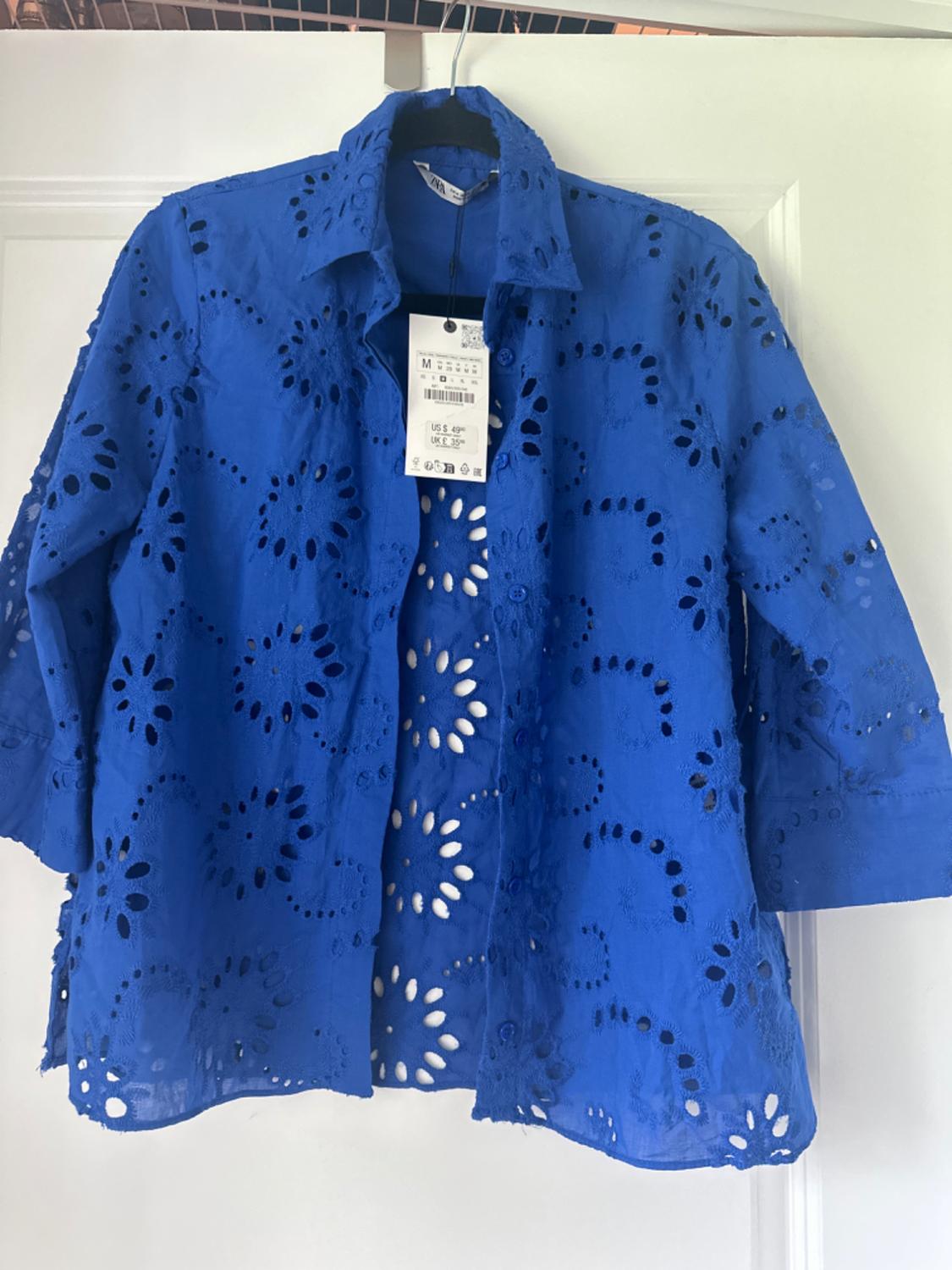 Front view of a blue blouse with openwork embroidery and a visible label on the collar.