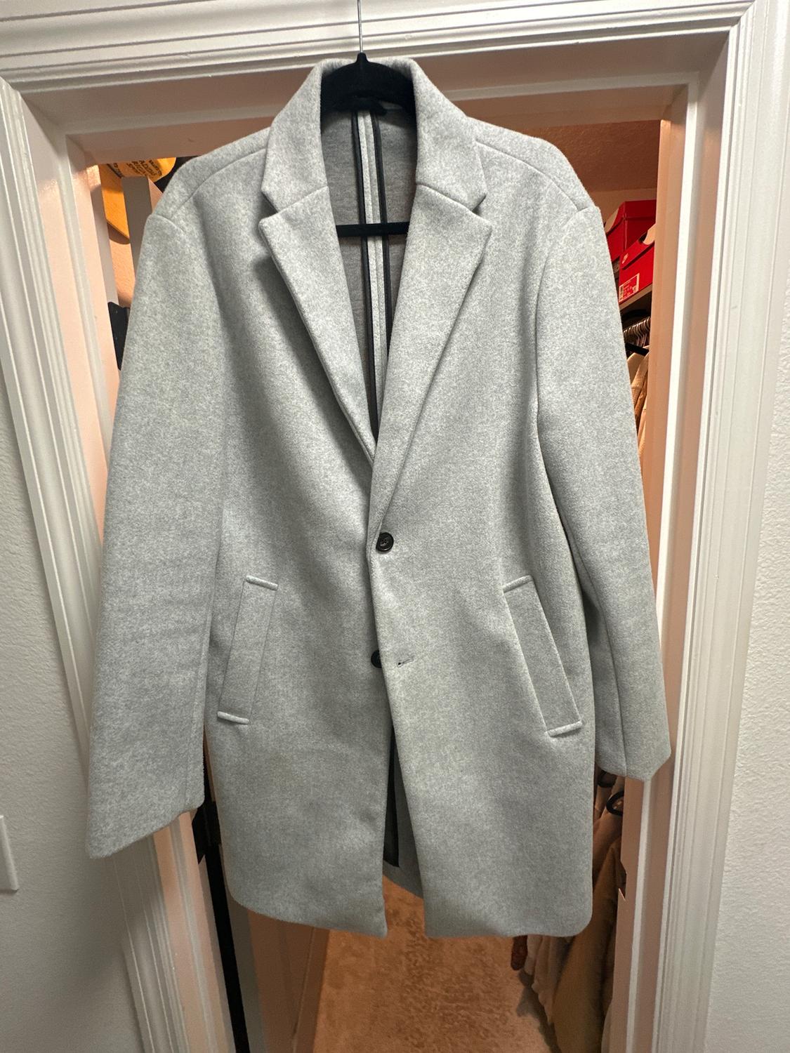 Light gray coat hanging on a hanger inside a door frame. It has lapels, two pockets, and one visible button.