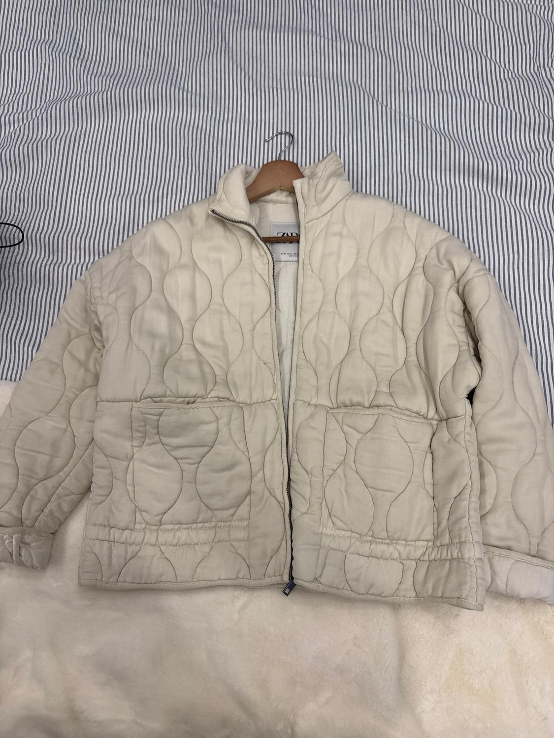 Beige quilted jacket with a wavy design, hanging on a hanger over a white and grey striped fabric.