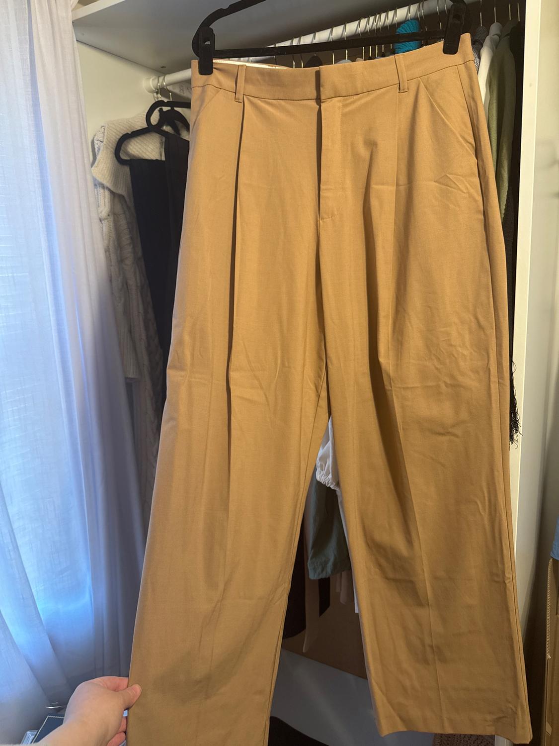 Some beige trousers are hanging on a hanger inside a wardrobe, whilst a hand is holding them. In the background, more hanging clothes and a white curtain can be seen.