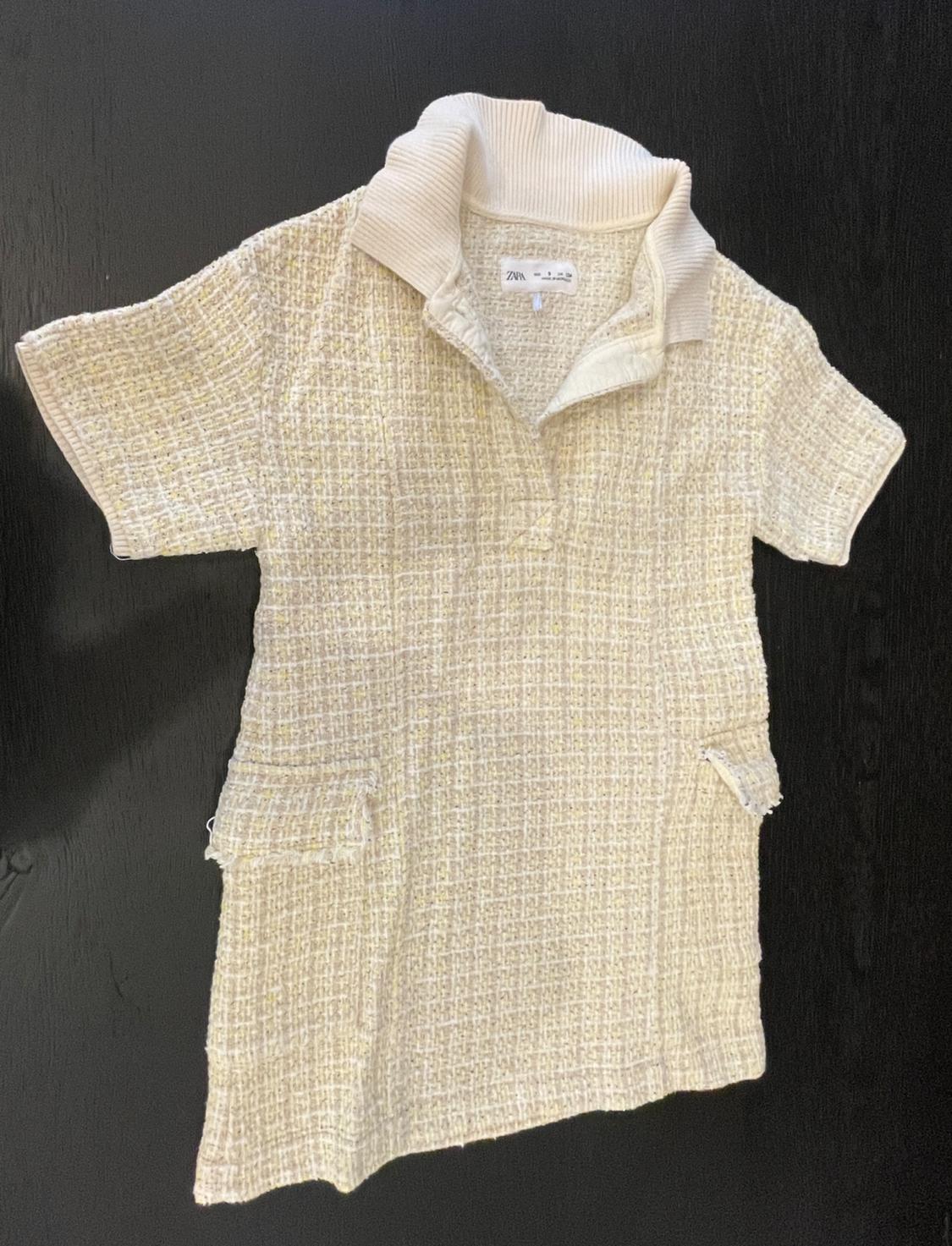 Beige short dress in a textured fabric with a polo collar and fringed pockets. ZARA label. Flat lay on a dark background.