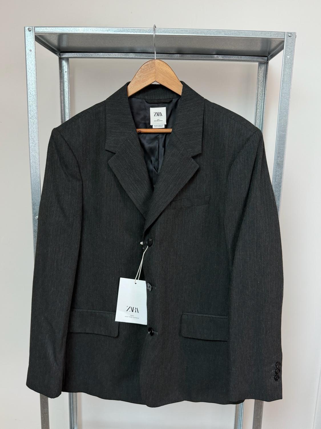 Dark grey suit jacket hanging on a wooden hanger on a metal coat rack. Visible labels with the text ZARA.