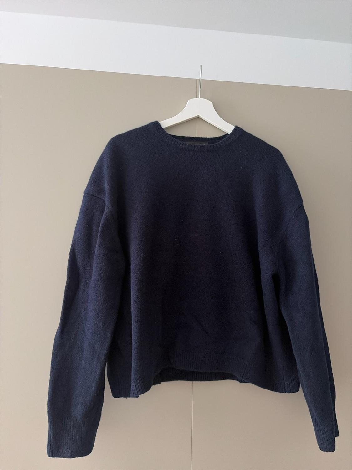 Navy blue oversized crew-neck knitted jumper, hanging on a white hanger against a plain beige wall.
