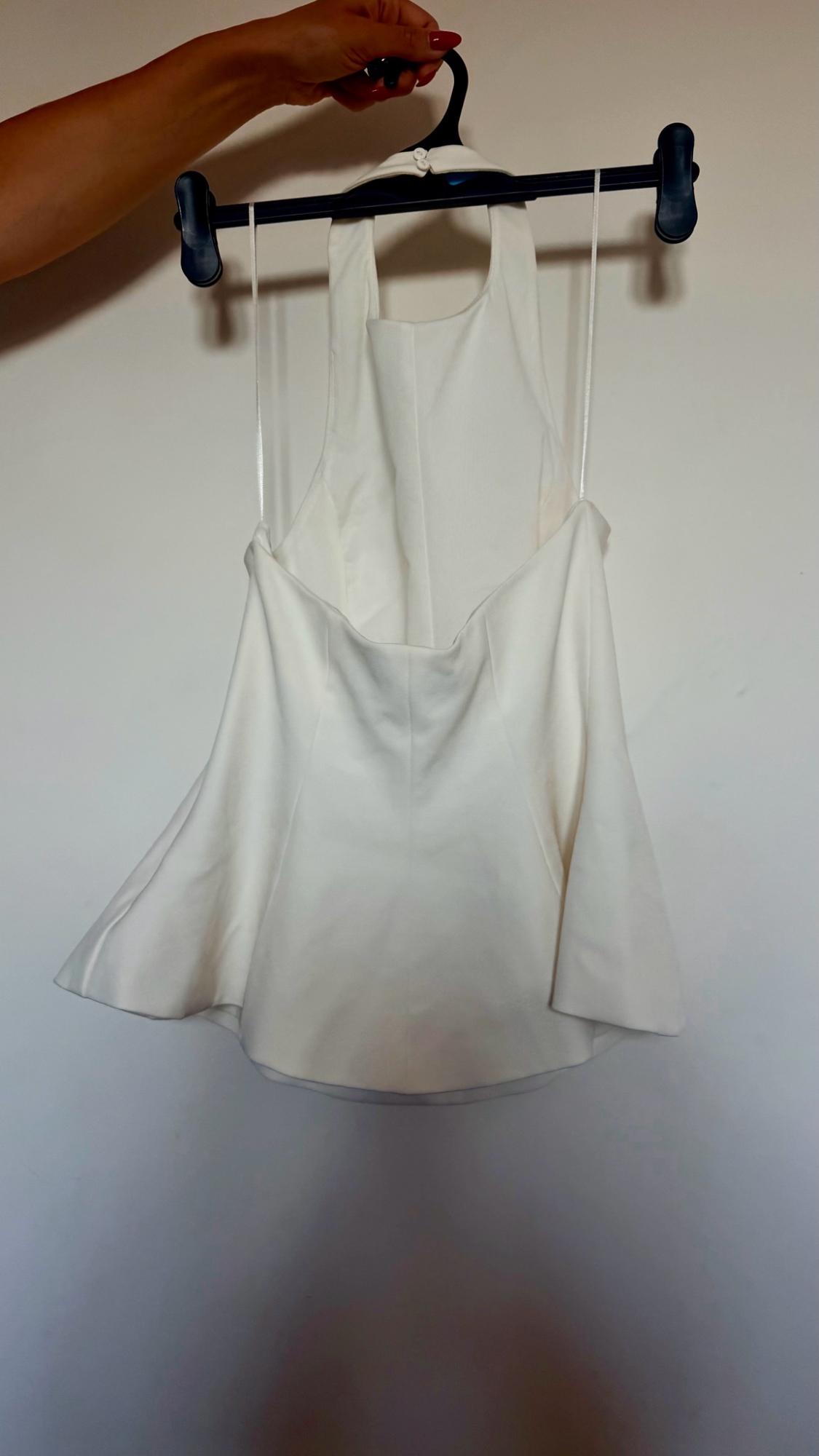 Arm holding a black hanger with a white halter-neck top with a flared hem against a white wall. Casual style.